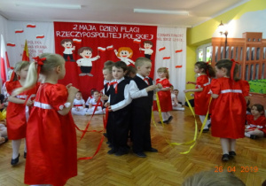 W środku koła stoją ubrani na galowo na chłopcy, na zewnątrz dziewczynki w czerwonych sukienkach. Dzieci tańczą ze wstążkami.
